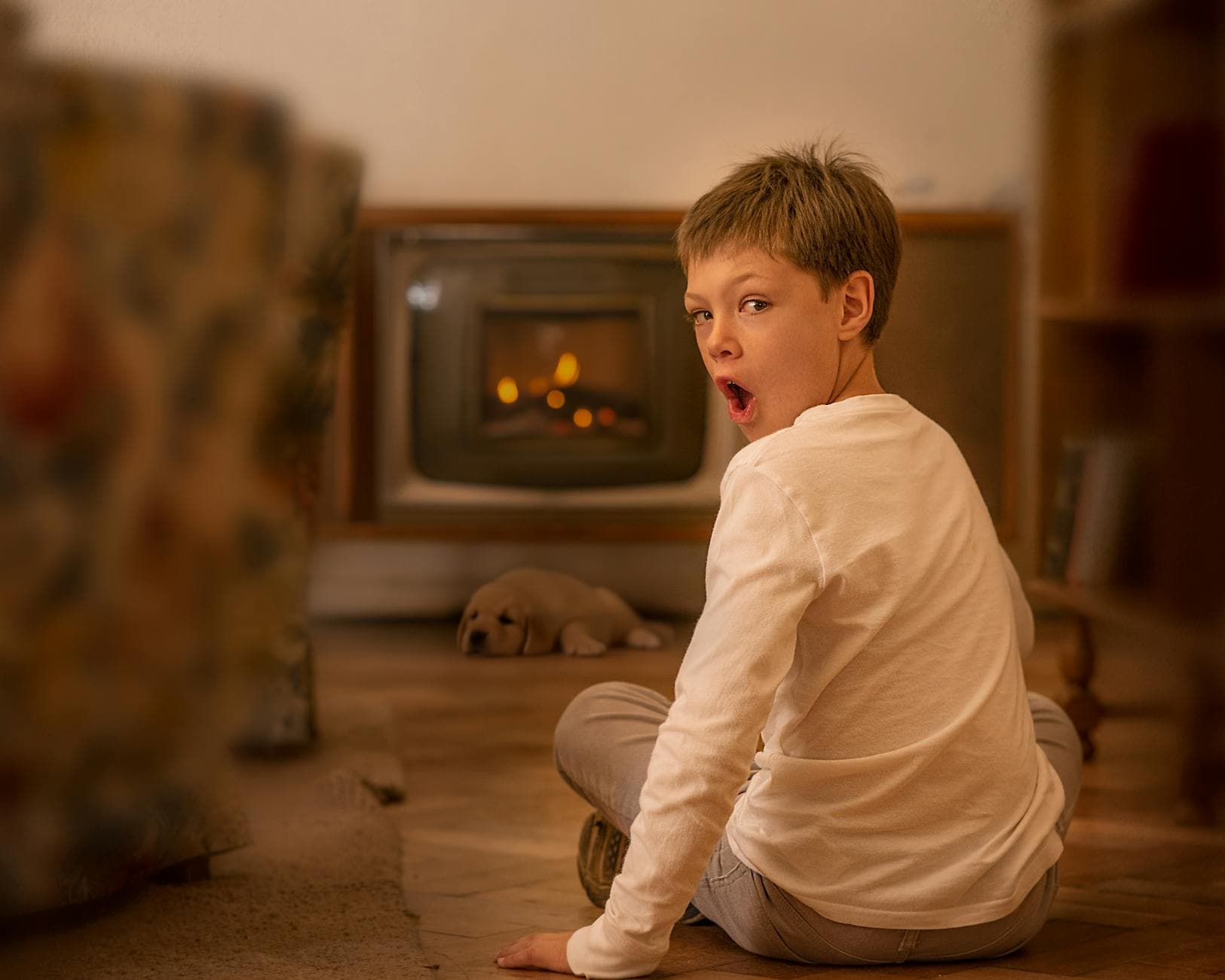 A child in a cozy indoor setting looks surprised while sitting by a warm fireplace.